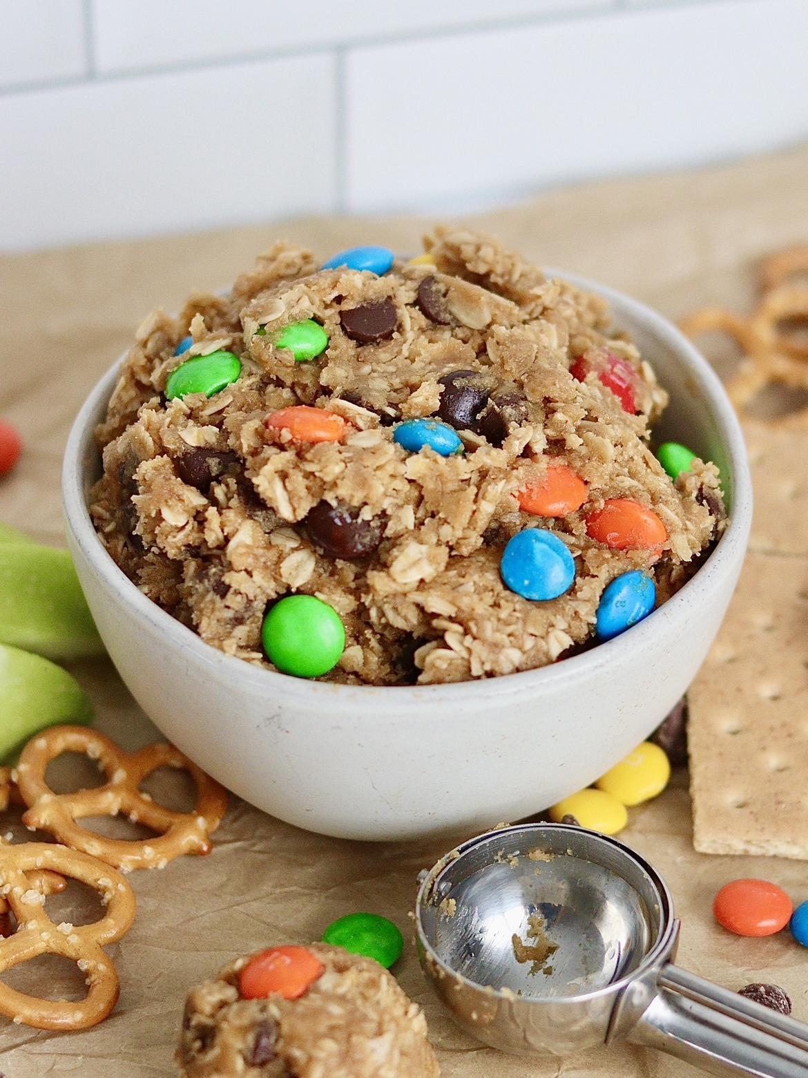 edible monster cookie dough in a bowl surrounded by apples, pretzels and graham crackers. A scoop of monster cookie dough is next to the bowl, with an empty cookie dough scoop