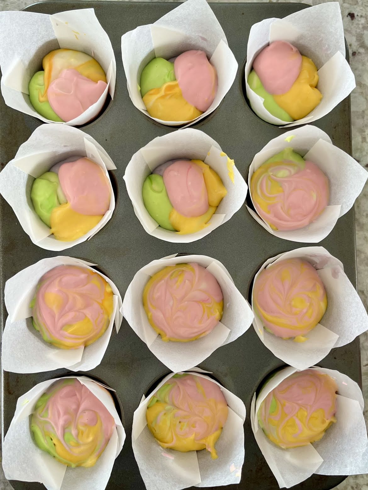 A cupcake tin filled with colored cheesecake batter, showing some that have been swirled and some that have not.