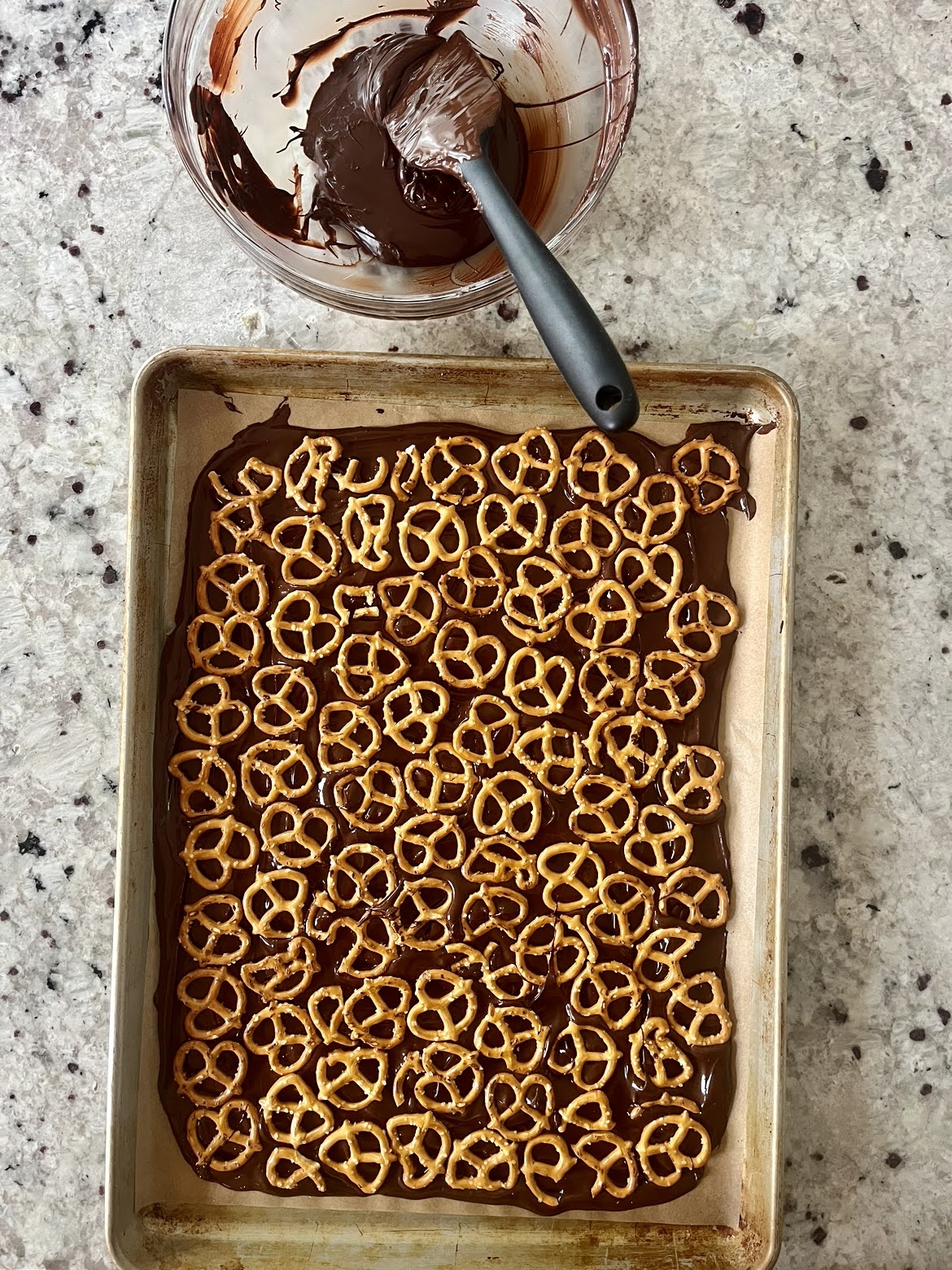 a process photo with melted chocolate spread throughout a cookie sheet with pretzels evenly placed on top