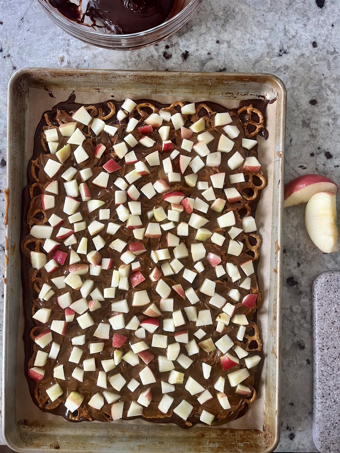 a process photo of melted chocolate, pretzels, caramel and chopped apples on a cookie sheet