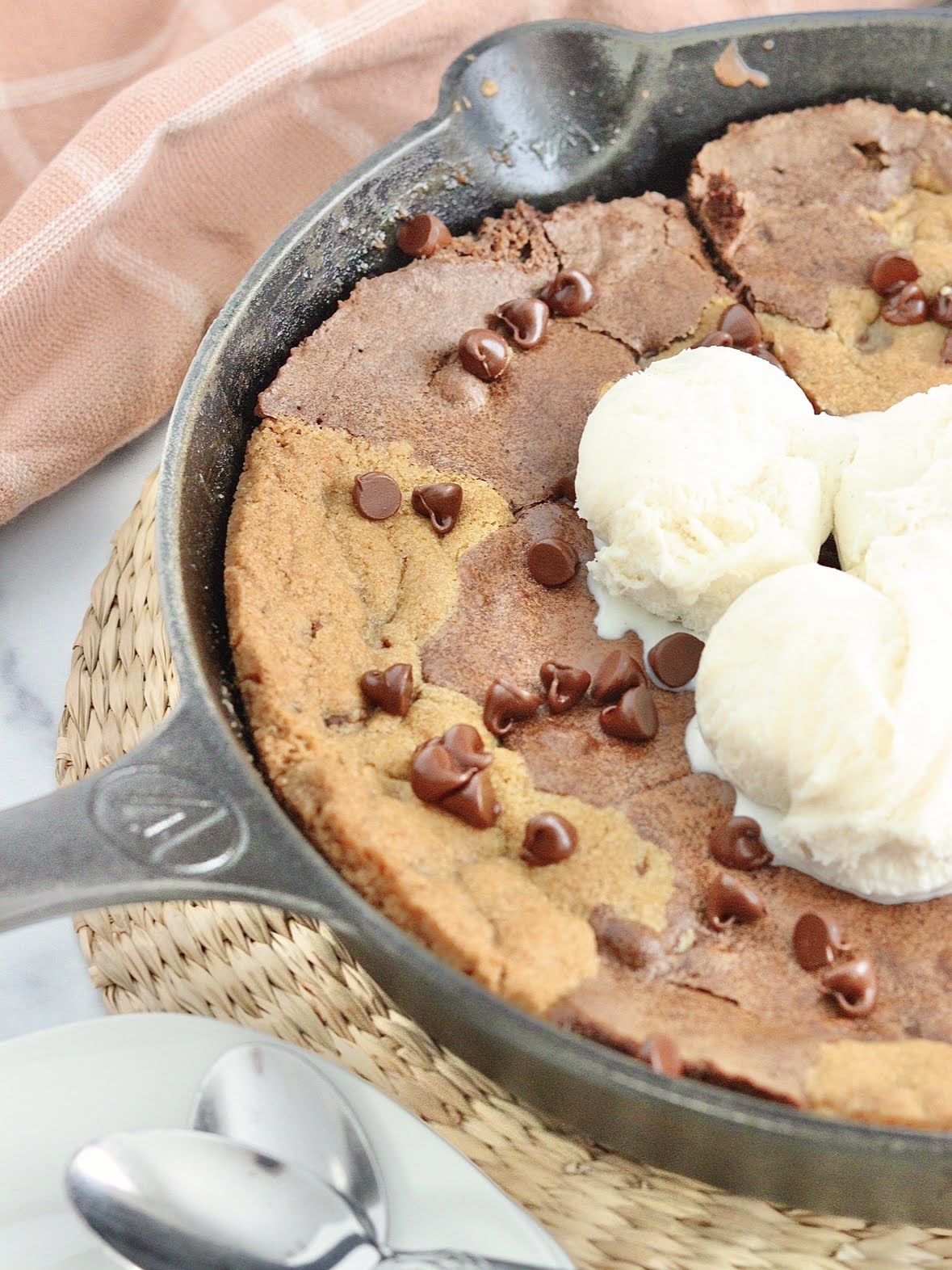 Photo of a cast iron skillet on a straw table mat filled with baked brownies and chocolate chip cookies topped with melting ice cream.