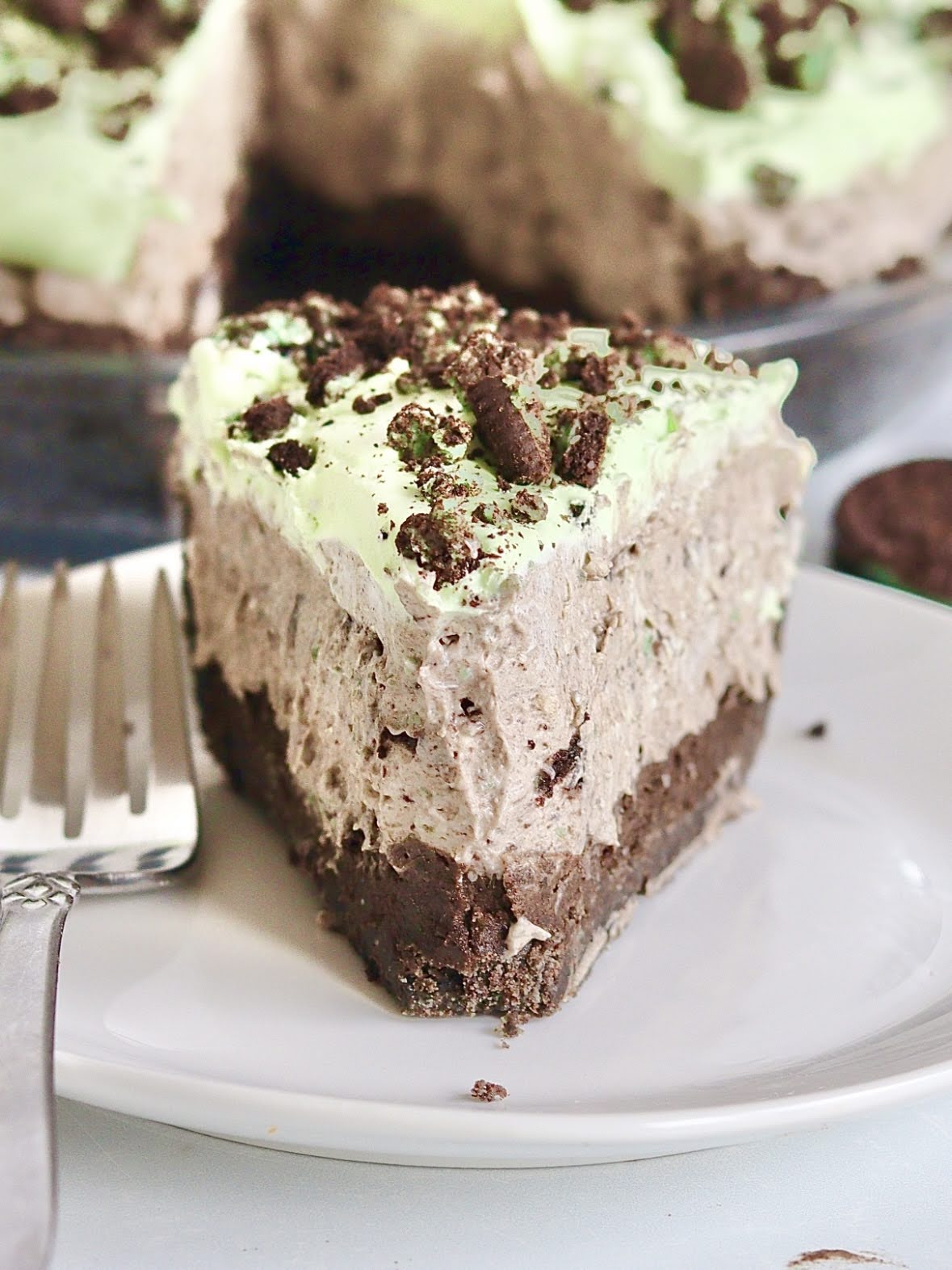 A close up photo of a plated slice of mint oreo truffle cream pie, showing the layers of crust, Oreo Truffle and Oreo cream layers, topped with cream whipped cream.